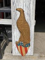 Front view of the full large cast metal American eagle wall plaque mounted on weathered wooden background, showing gold-colored eagle perched on a patriotic shield with red and white stripes and blue with stars.