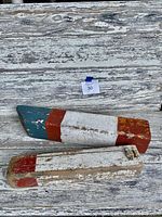 Two antique wooden buoys laying on weathered white wooden surface showing full bodies with distinct red, white, and blue paint.