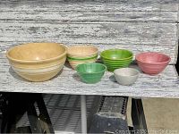 Front view of six antique stoneware bowls in various colors and sizes arranged side by side on a weathered wooden surface.