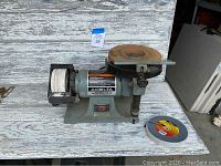 Front perspective of Delta sharpening center showing buffing wheel at left, motor center, and grinding wheel with rust and plastic guard at right. Extra cutting/grinding disc lies beside machine.