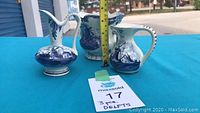 Three-piece Delftware pottery set on a blue table: tall narrow-necked pitcher, wide-mouthed scalloped pitcher, and smaller rounded pitcher with blue and white windmill designs.
