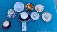 Overview of assorted tea cups and saucers on blue cloth including blue and gold striped set and various white cups with floral and gold details