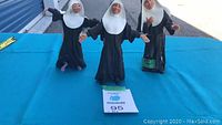 Three nun figurines standing on blue surface outside a storage facility, showing frontal view of the lot