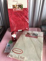 Two tablecloths in original packaging and Santa carving figurine on pink surface with gray corrugated background.