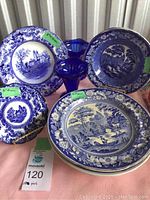 Full view of the blue and white plates of varying sizes with two blue glass sundae dishes, showing detailed scenic patterns and floral borders.