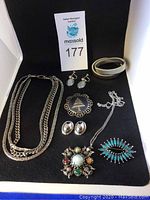 Photo showing the assortment of vintage jewelry on black background including signed Jacobite multi-strand necklace, Christmas tree brooch, tri-silver bracelet, clip-on earrings, colorful brooches, and pendant necklace with blue stone earrings.