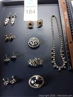 Full view of all jewelry items arranged on black surface with ruler for scale shows five pair of rhinestone earrings, three pins including a thistle sterling silver pin, and a rhinestone necklace.