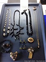 Photo showing four necklaces with black beads, a gold-tone pendant necklace, a gold-tone men's ring with diamond-like stones, and various brooches and earrings arranged on a tray.