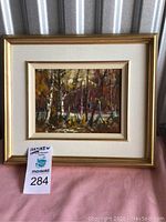 Framed painting of a birch tree forest scene in a gold frame sitting on pink fabric.