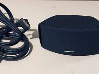 Side view of black Bose fabric speaker with multi-pin connector connection cable coiled beside it.