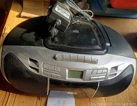 Black RCA boombox radio with power adapter on top, front speaker grille, control buttons, and small digital display.