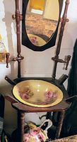 Vintage wooden wash stand with an oval mirror, brass faucets, and a yellow porcelain basin with pink rose design visible. The basin has no chips. Missing towel holders.