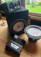 Photo showing Alpine M350 power amplifier with cables, large subwoofer speaker in black carpeted enclosure, loose subwoofer speaker, and 3-driver speaker unit with black enclosure.