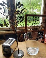 Two metal decorations including a standing leaf branch and a framed floral vase wall piece, stainless steel toaster, and clear glass bucket on a table near a window.