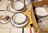 Photo of multiple place settings stacked and laid out showing plates, bowls, cups, saucers, a large serving bowl, and a platter with a blue band and floral pattern around the edges.