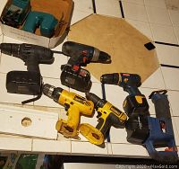 Seven cordless drills and one reciprocating saw displayed on a counter, showing condition and brands.