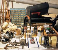 Photo showing parts bin with small drawers, spray paint cans, paint tubes, mailbox, toolboxes, wheels, hand grinder, and other hardware items arranged on white tiled surface.