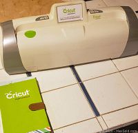 View of Cricut Expression 2 cutting machine showing its white body with grey ends and accompanying green manual on a tile surface.