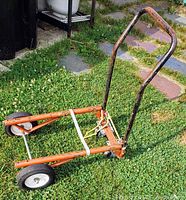 Rusty orange dolly with three wheels standing on grass, bent handle section visible