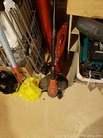 Cordless string trimmer standing upright beside a red Bosch metal toolbox and other assorted tools on a floor.