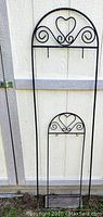 Two wrought iron garden decoration pieces leaning against a shed, showing height and heart-shaped decorative elements.