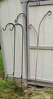 Four large metal shepherd's hooks of varying designs leaning against a shed wall outdoors on grass.