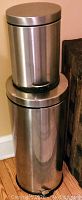 Two stainless steel trash cans, one small and one large, stacked vertically and positioned against a wall and wooden floor
