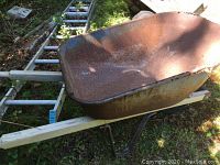 Aluminum extension ladder laid out on grass next to a rusty metal wheelbarrow with signs of weathering and wear.