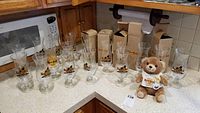 Collection of 13 Hard Rock Cafe hurricane glasses and one stuffed bear on a kitchen counter; some glasses in packaging.