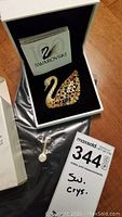 Swarovski swan brooch inside black velvet box with certificate and packaging visible, alongside maxsold lot label and small key charm