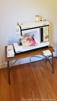 Kenmore white sewing machine sitting on small wooden sewing table on hardwood floor against wall, with power cord and instruction manual placed nearby.