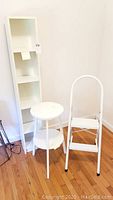 Photo showing white tall shelf unit, white round table, and white step stool on wooden floor against white wall.