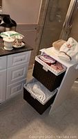 White freestanding storage unit with two woven baskets containing towels and bath sponge in bathroom setting.