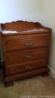 Full front view of wooden night stand with scalloped back edge and three drawers.