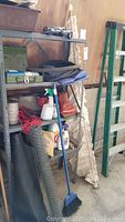 Photo of a gray metal shelf with various paint supplies, cleaning products, garden hoses, tools, chicken wire, and rope. Orange rain boots and a blue broom lean against the shelf, with a green step ladder to the right.