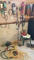 Green Craftsman electric hedge trimmer lying on the floor with yellow extension cord, red and gray weed wacker standing against the wall, belts hanging on pegboard, and hand tools nearby.