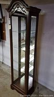 Front and side view of a tall wooden curio cabinet with glass panels and shelves, showing carved decorative details on top.