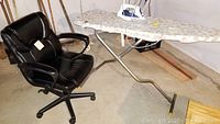 Photo showing black office chair next to ironing board with iron on top in basement setting.