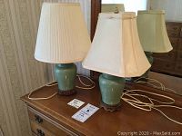 Two celadon ceramic lamps with fabric shades and carved wooden bases shown side by side on a wooden surface.