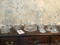Full lot of crystal pieces on wooden surface showing various sizes and shapes, including a covered dish, a bowl, stemmed glasses, and small bowl with pointed lid.