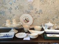 Fine china tableware displayed on a dark wood surface with floral wallpaper background. Includes cake stand with candle holders, floral plate, various small bowls, rectangular blue and white plate, hexagonal teapot, and several trays.