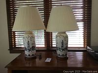 Pair of Asian vase style table lamps on a wooden surface near window blinds. Both lamps have cream ceramic bases with indigo and rust painted designs and pleated cream lamp shades.