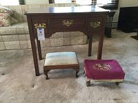 Front view of pine hall table with three drawers showing brass hardware, two small footstools in front, one with white upholstery and one with reddish-purple needlepoint fabric.