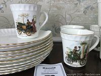Stack of white scalloped snack plates with gold trim, with two vintage automobile print mugs on top of the stack.