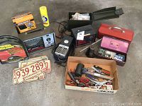 Photo showing Craftsman Engine Analyzer, vintage license plates, assorted hand tools, battery charger, inflator, and several boxes including ammo box and tool boxes.