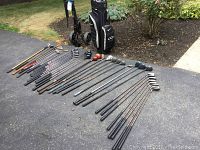 Full set of various golf clubs laid out on driveway showing wood and metal club heads and shafts.