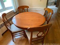 Full view of round maple table and four matching chairs around it on wood floor near window.