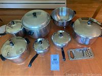 Overview of all pots, pans, colander, and cheese grater arranged on wood table with product card
