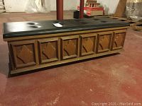 Full profile view showing chest with black tufted Naugahyde top, six decorative wood panels on front, and wheeled base.