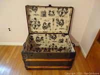 Photo of trunk open to show Victorian style printed paper lining inside lid and base, featuring portraits and decorative elements in black and white.